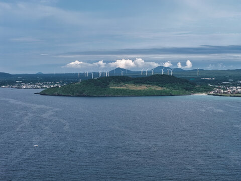Aerial view of a lush green coastal hill surrounded by the blue ocean with wind turbines on the distant hills under a cloudy sky in South Korea.
