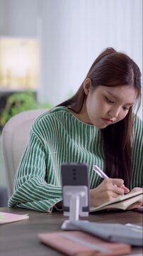 Young Asian student checking smartphone while studying with textbooks and notes at home desk