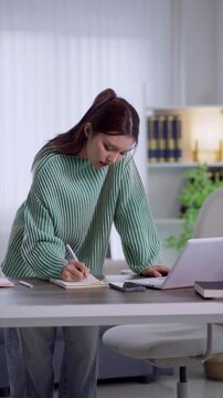 Young Asian woman writing notes and using laptop for remote work at home office desk