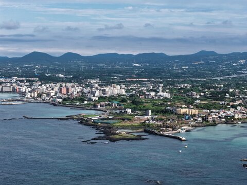 Aerial view of a coastal town on Jeju Island featuring basalt rock formations, turquoise waters, and residential buildings under a clear sky in South Korea.
