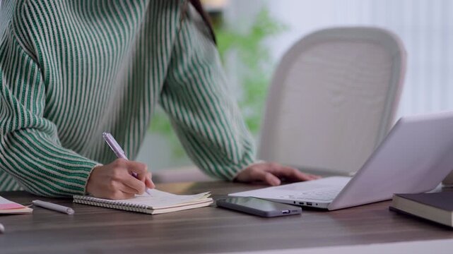 Asian woman writing notes in notebook while using laptop for online learning at home office desk