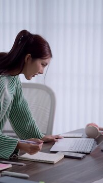 Young Asian woman writing notes in notebook while using laptop for remote work at home desk with copy space