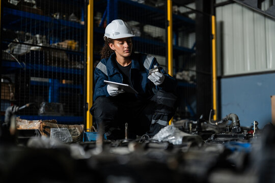 Female engineer in hard hat using tablet to inspect e-waste and old machinery parts in a warehouse for Industry 4.0 circular economy, sustainable recycling, and inventory management.