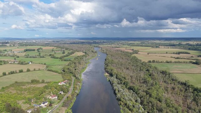 The Loire River winding through the rural landscape of France. The scene features lush green fields, patches of forest, scattered country houses, and dramatic clouds in the sky, aerial view.