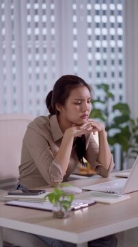 Young Asian professional woman looking thoughtfully away while sitting at laptop desk in modern home office