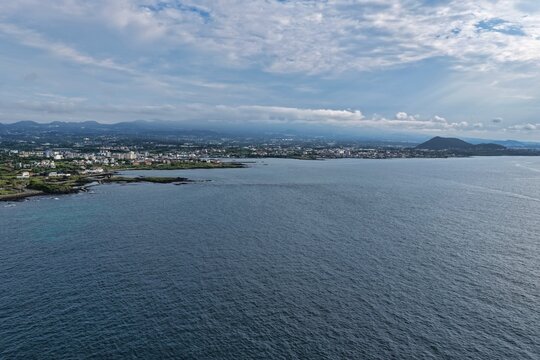 Aerial view of the coastline of Jeju Island with a volcanic cone in the background, coastal town buildings, rocky shore, and deep blue sea in South Korea.