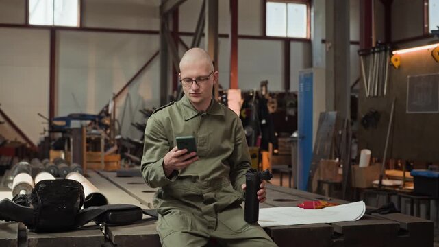 Young adult Caucasian male welder sitting in workshop, drinking from thermos and using smartphone. Clip is perfect for industrial manufacturing marketing or workforce recruitment campaigns