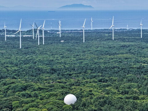 Aerial view of a wind farm with multiple turbines situated in a dense forest, featuring a large white radar dome in the foreground and the ocean in the distance in South Korea.