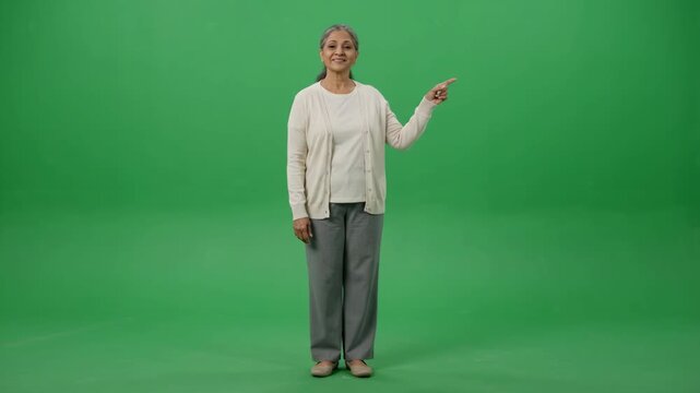 Smiling senior Indian woman pointing to the right on a green screen background, full body shot.