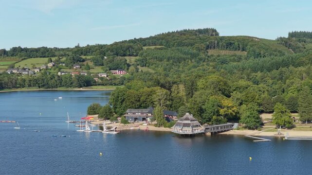 Lac des Settons in Morvan, France, showing a lakeside marina with sailboats, a unique waterfront building, sandy beach, and dense green forest, aerial view.