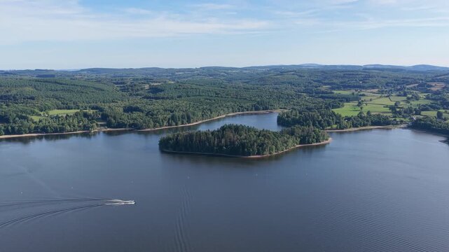 Lac des Settons in the Morvan region of France. The clip showcases a forested island surrounded by calm lake waters, with a boat creating ripples on the surface, aerial view.