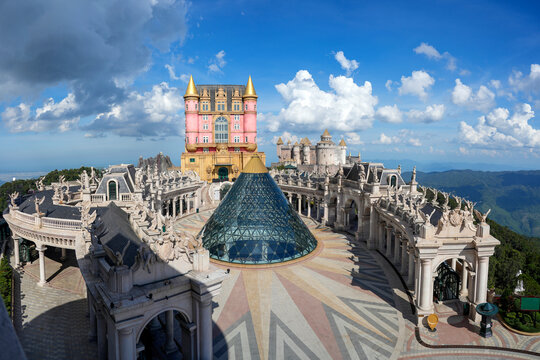 Brightly colored castle stands proud amidst intricate architecture, surrounded by lush mountains hills and fluffy clouds during a sunny afternoon