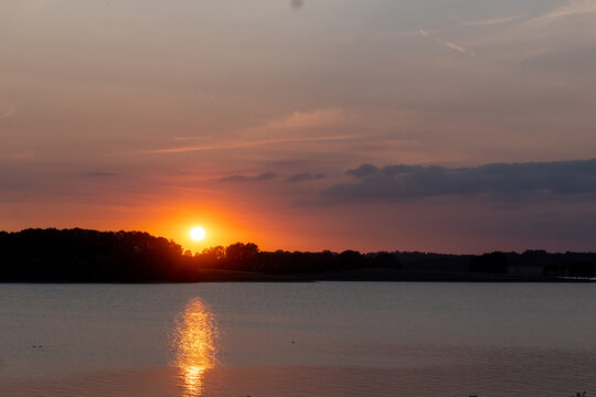 Orange sun setting over calm water with a golden reflection and silhouetted shoreline. Rutland Water, Rutland, England, UK