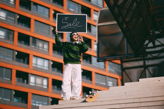 Young person standing on concrete stairs in a modern city setting, holding a blackboard with the word sale written on it, and shouting, actively promoting a discount or special offer