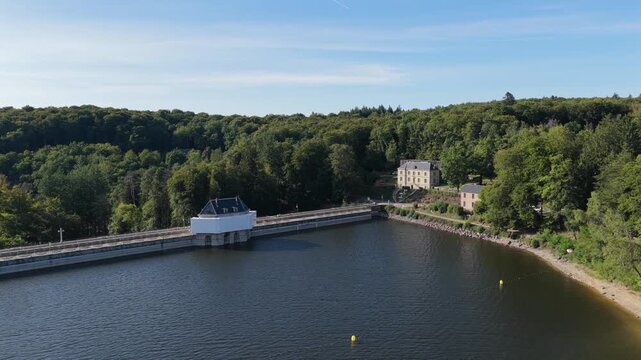 The Lac des Settons dam in Morvan, France, showing the lake, the dam structure, and surrounding dense green forest under a clear blue sky, aerial view.