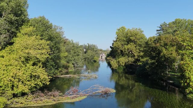 A tranquil view of the Canal du Loing in Moret-sur-Loing, France, captured on a clear sunny day. The scene features calm water reflecting dense green trees lining both banks of the canal.