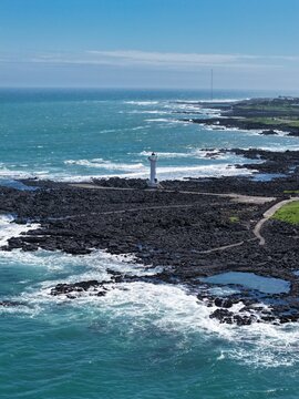 Aerial view of a white lighthouse standing on a black volcanic rocky coastline with crashing ocean waves under a clear blue sky in South Korea.