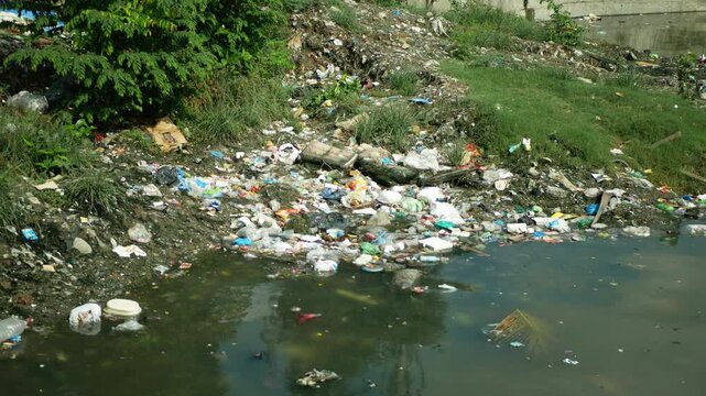 Heavily polluted river waterway pollution garbage trash surrounded vegetation contamination plastic bottles, plastic bags, food packaging, containers, solid waste floating river water scattered muddy