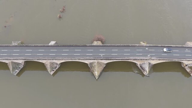 A medieval stone bridge in Gien, France, spanning a wide river. The video shows vehicles driving across the historic bridge, highlighting its stone arches and geometric structure, aerial view.