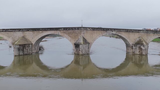 A historic medieval stone bridge spanning a river in Gien, France. The bridge features multiple large arches and is reflected in the calm water below.