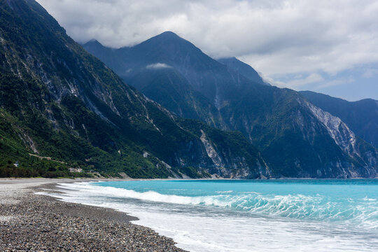 Scenic Qingshui Cliff in Hualien, Taiwan. High coastal mountains meet the turquoise Pacific Ocean. Pebble beach with breaking waves under overcast sky.