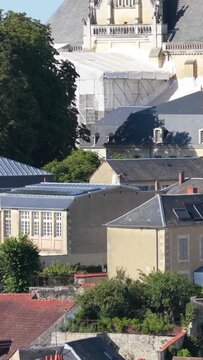 The upper portion of a historic church under renovation, with scaffolding and white tarps visible on the roof, surrounded by classic French architecture and leafy trees, vertical 9:16 view.