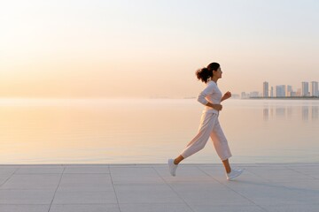 Fototapeta premium Woman running along a calm seaside promenade at sunrise with city skyline in the distance for wellness background and active lifestyle banner design