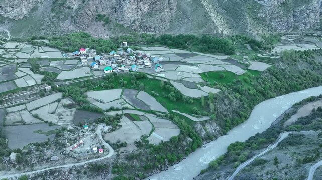 Aerial top view of riverside village in mountain with colorful houses and farm terraces in the Lahaul Valley shot by a drone flying towards the village along a river
