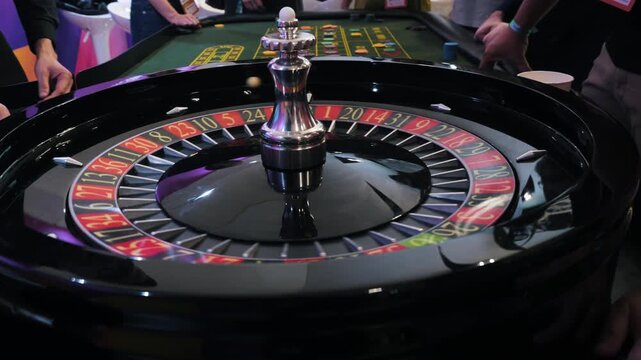 Spinning casino roulette wheel with glossy black rim, numbered betting pockets, chrome spindle, blurred ball movement, and surrounding hands in colorful indoor gaming atmosphere