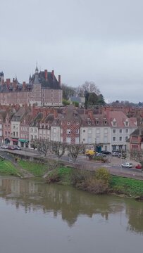 Gien city center in France, featuring a scenic view of the historic Chateau de Gien, traditional French townhouses, and the Loire river in the foreground, vertical 9:16 view.