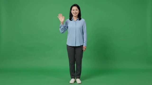 Full body shot of a smiling Asian woman waving hello on a green screen background, expressing a friendly greeting.