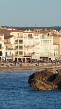 Vertical 9:16 video showing a lively seafront promenade in L'Escala, Spain. A large rocky outcrop is visible in the foreground, and calm blue sea water fills the lower part of the frame.