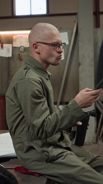 Young adult Caucasian male welder sitting in workshop, drinking from thermos flask and using smartphone. Footage is perfect for blue-collar recruitment or industrial app promotions, vertical