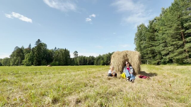 A mother and son sit and rest by a large haystack in a sunny meadow. They feel happy, peaceful, and relaxed during this joyful outdoor break under a blue sky.