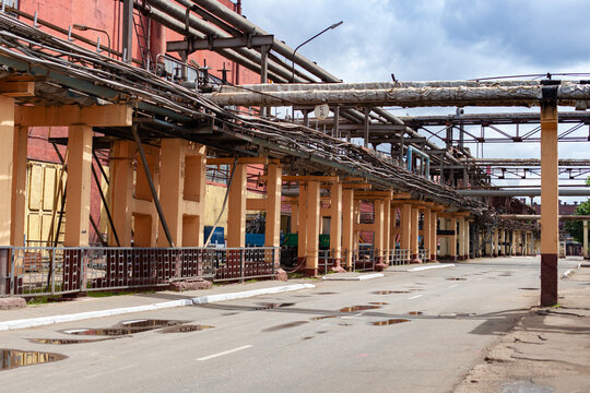 Industrial landscape featuring  factory exterior with complex piping systems, metal structures, and asphalt road with puddles. Industrial infrastructure, heavy manufacturing, engineering.