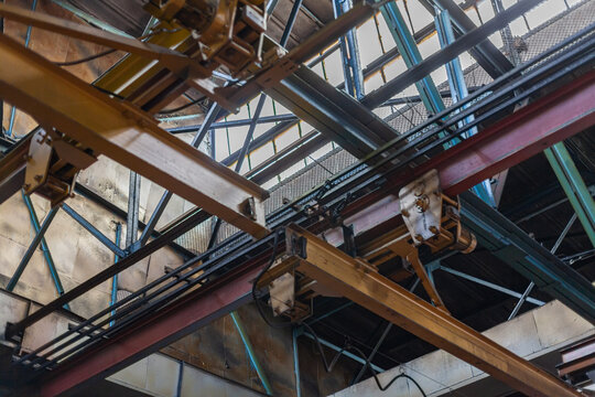 Low-angle view of industrial ceiling structure with overhead cranes and steel beams inside tractor manufacturing plant. Industrial interior, heavy industry, construction, mechanical infrastructure.
