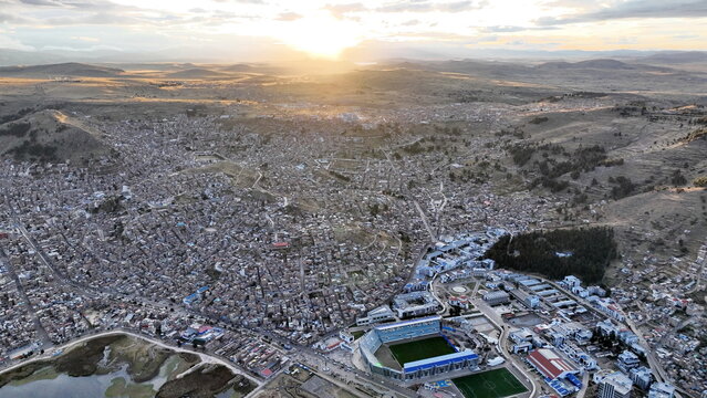 Aerial view of Puno city on the shores of Lake Titicaca, featuring the Enrique Torres Belon stadium, dense urban housing, and rolling hills under a golden sunset sky in Puno, Puno, Peru.