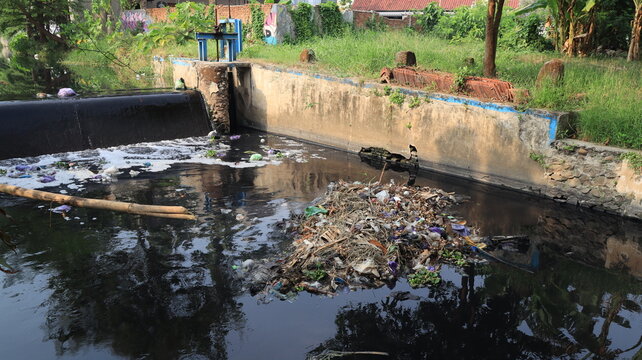 A residential canal carries visibly polluted dark water and floating plastic, bordered by concrete walls, trees, and houses in a densely developed neighborhood.