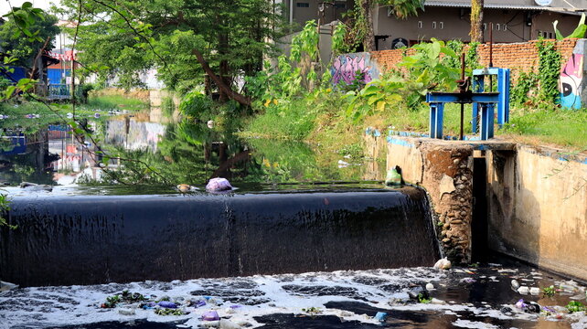 Dark wastewater flows through concrete neighborhood canal, carrying rubbish while homes, tree, and drainage structures frame the polluted urban landscape. also.