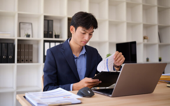 Young Asian businessman reviewing reports while working on a laptop in the office.