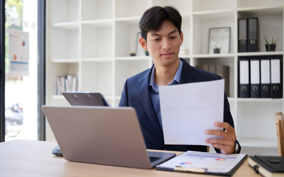 Young Asian businessman reviewing reports while working on a laptop in the office.