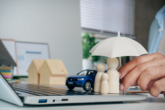 Hand holding small umbrella protecting wooden peg doll family and toy car on laptop keyboard with wooden house models in background, concept of comprehensive insurance for home, car, and family.