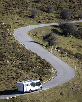 View of a motorhome driving along a winding mountain road near the Lakes of Covadonga, surrounded by grassy hills and rocky terrain in Asturias, Spain.