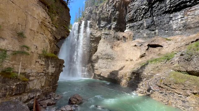 Johnston Creek river in Johnston Canyon in the Banff National Park. British Columbia, Canada. Waterfall flows over rocks into a clear pool, framed by cliffs and trees under a sunny sky