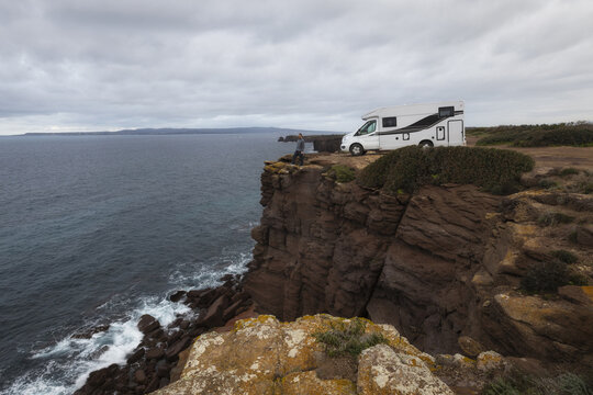 View of a camper van parked on the rugged red cliffs of Sant'Antioco overlooking the sea with a person standing near the edge under a cloudy sky in Sant'Antioco, Sardinia, Italy.