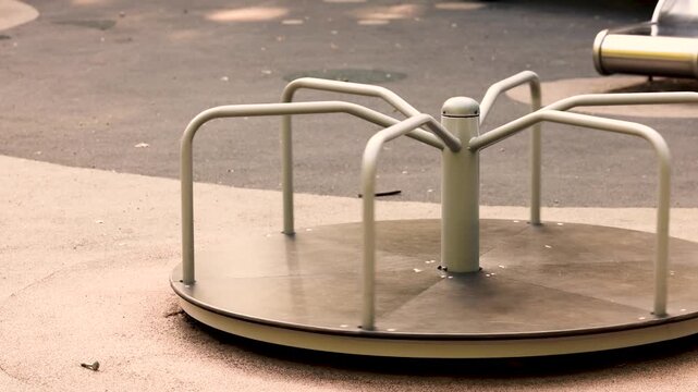 Empty round metal merry go round spins slowly on modern playground equipment, motion blur capturing smooth rotation, surrounded by wooden climbing structures and green rubber safety surface in park