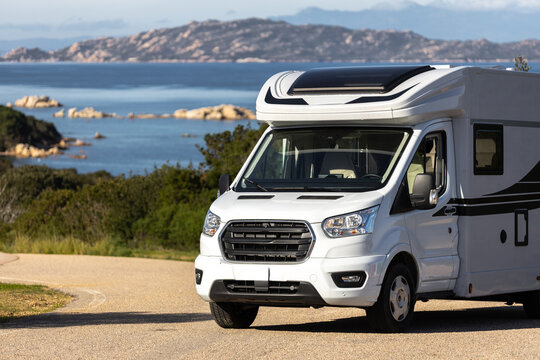 View of a white motorhome parked on a coastal road overlooking the blue sea and rocky islands in Palau, Sardinia, Italy.