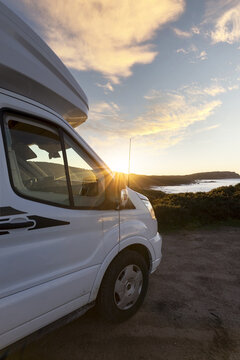View of a white camper van parked on a dirt path overlooking the coastline at sunset with golden light reflecting off the vehicle in Olbia, Sardinia, Italy.