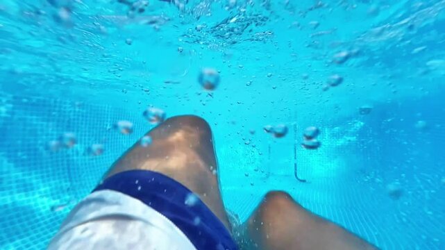 Point of view underwater view of a man kicking his legs and swimming in a bright blue swimming pool with water bubbles floating by. Summer vacation concept.
