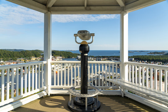 View of a coin-operated binocular viewer at a scenic lookout point overlooking the coastal town, with boats in the harbor, white wooden houses, and a blue sky in Mandal, Agder, Norway.